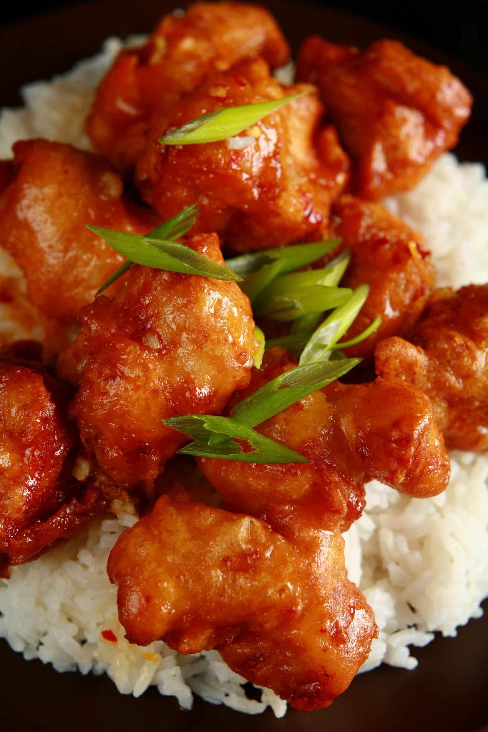 A big helping of gluten free orange chicken: Chunks of battered and deep fried chicken coated in a glossy orange sauce, garnished with sliced green onions, and served over a bed of rice.