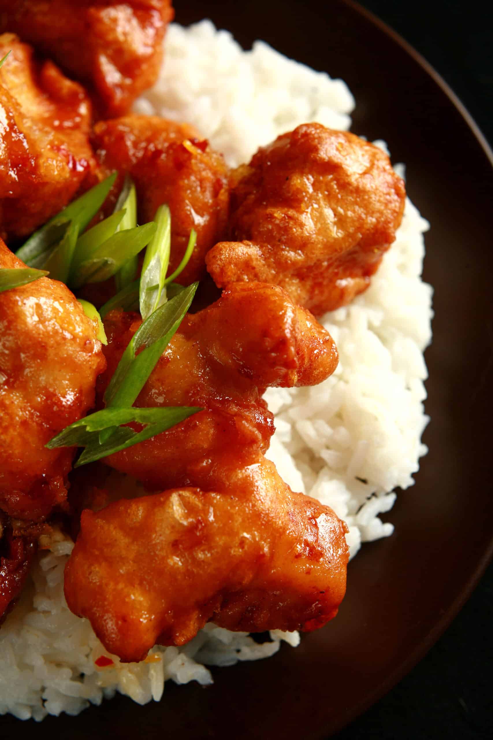 A big helping of gluten-free orange chicken: Chunks of battered and deep fried chicken coated in a glossy orange sauce, garnished with sliced green onions, and served over a bed of rice.