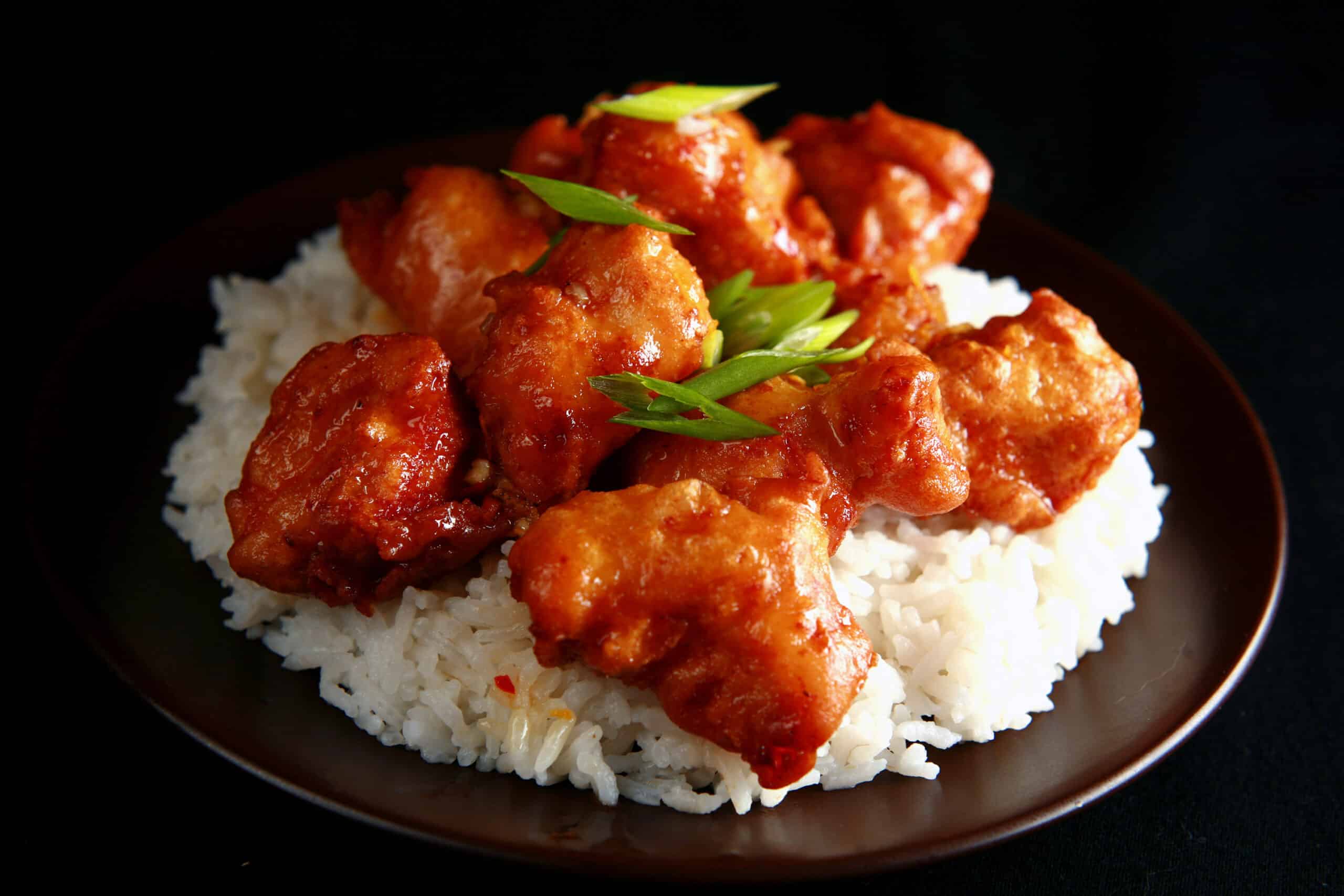 A big helping of gluten-free orange chicken: Chunks of battered and deep fried chicken coated in a glossy orange sauce, garnished with sliced green onions, and served over a bed of rice.