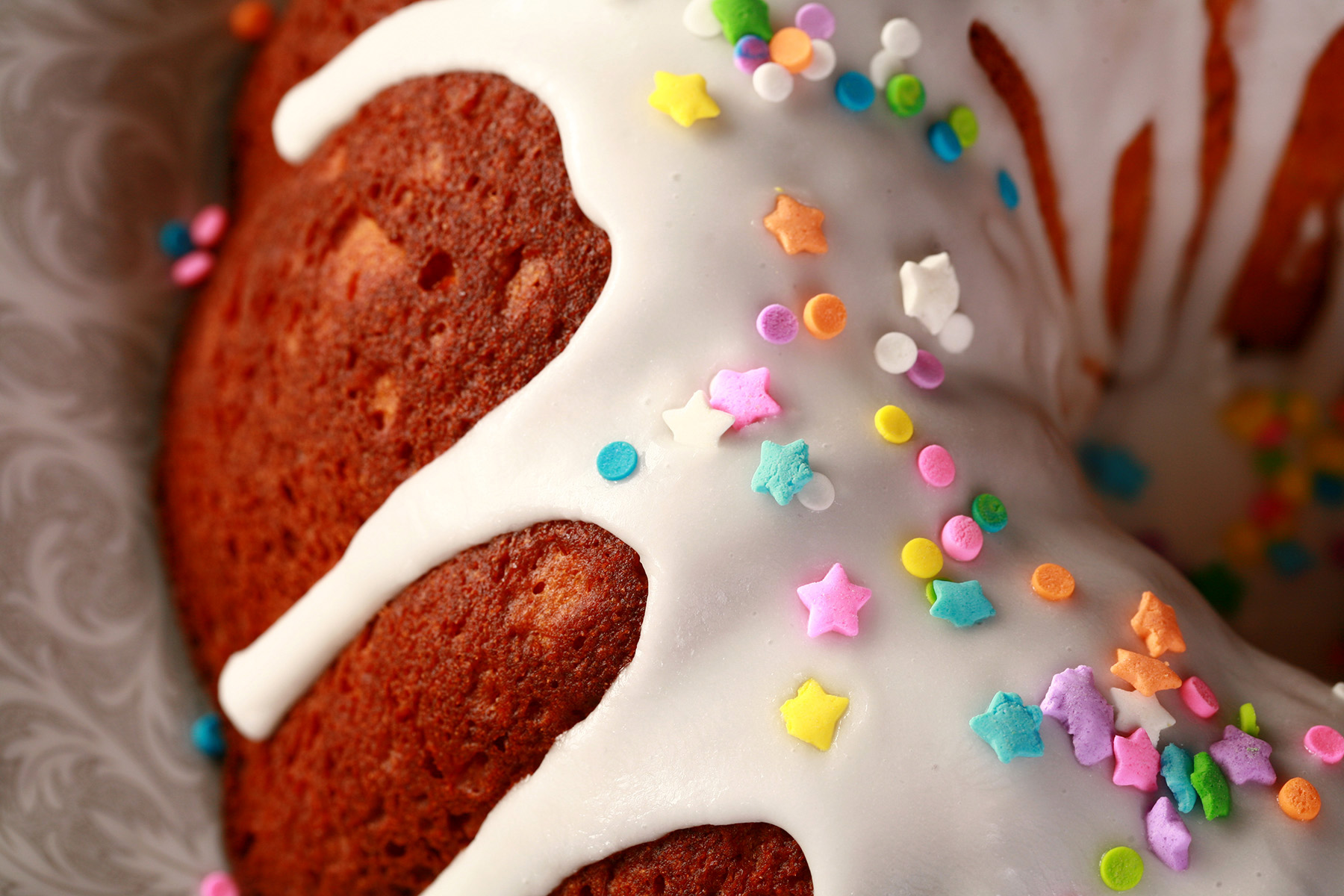 A close up view of a gluten-free paska. It's in the shape of a bundt pan, with dripping white glaze and topped with pastel coloured sprinkles.