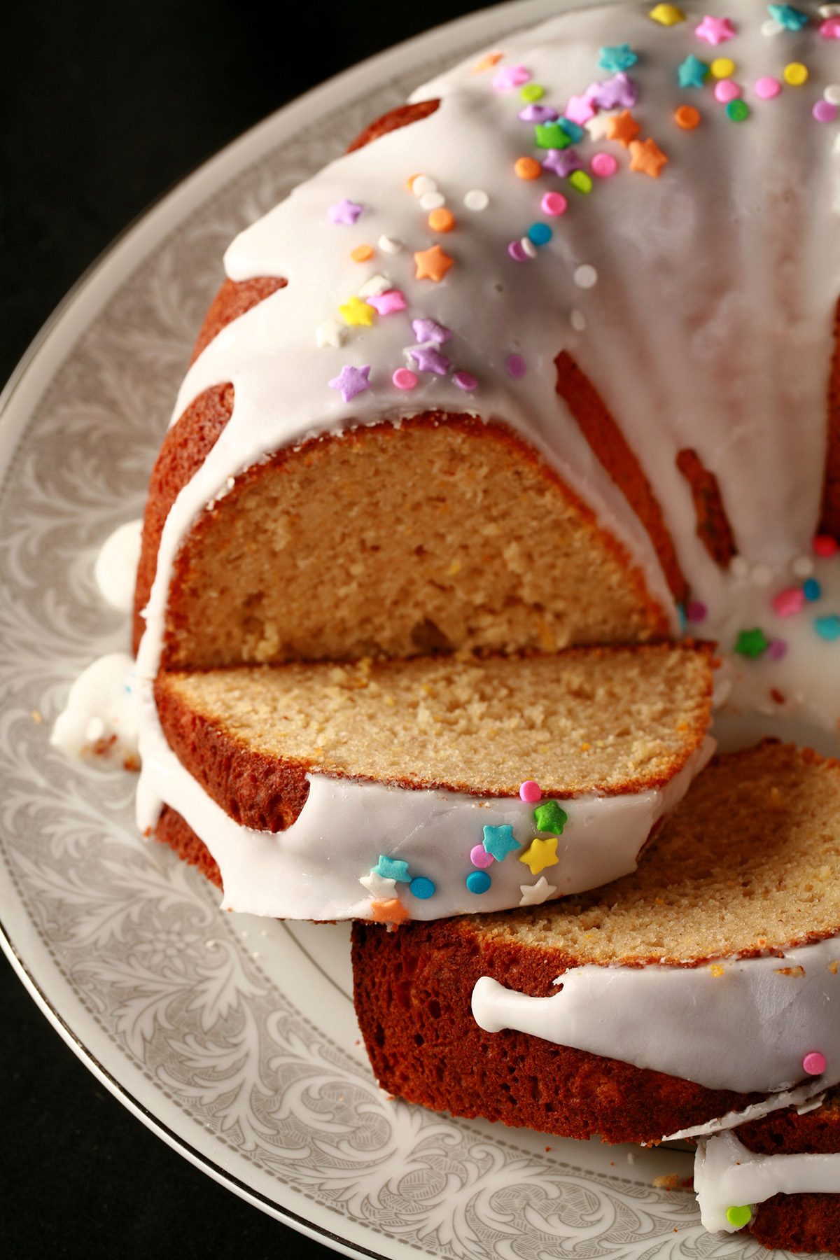 A close up view of a gluten-free paska. It's in the shape of a bundt pan, with dripping white glaze and topped with pastel coloured sprinkles. A section has been sliced, the slices are fanned out on the plate.