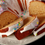 A close up view of a gluten-free paska. It's in the shape of a bundt pan, with dripping white glaze and topped with pastel coloured sprinkles. A section has been sliced, the slices are fanned out on the plate.