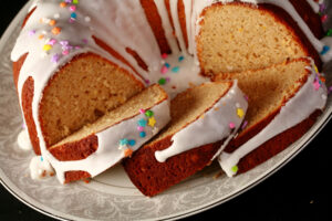 A close up view of a gluten-free paska. It's in the shape of a bundt pan, with dripping white glaze and topped with pastel coloured sprinkles. A section has been sliced, the slices are fanned out on the plate.