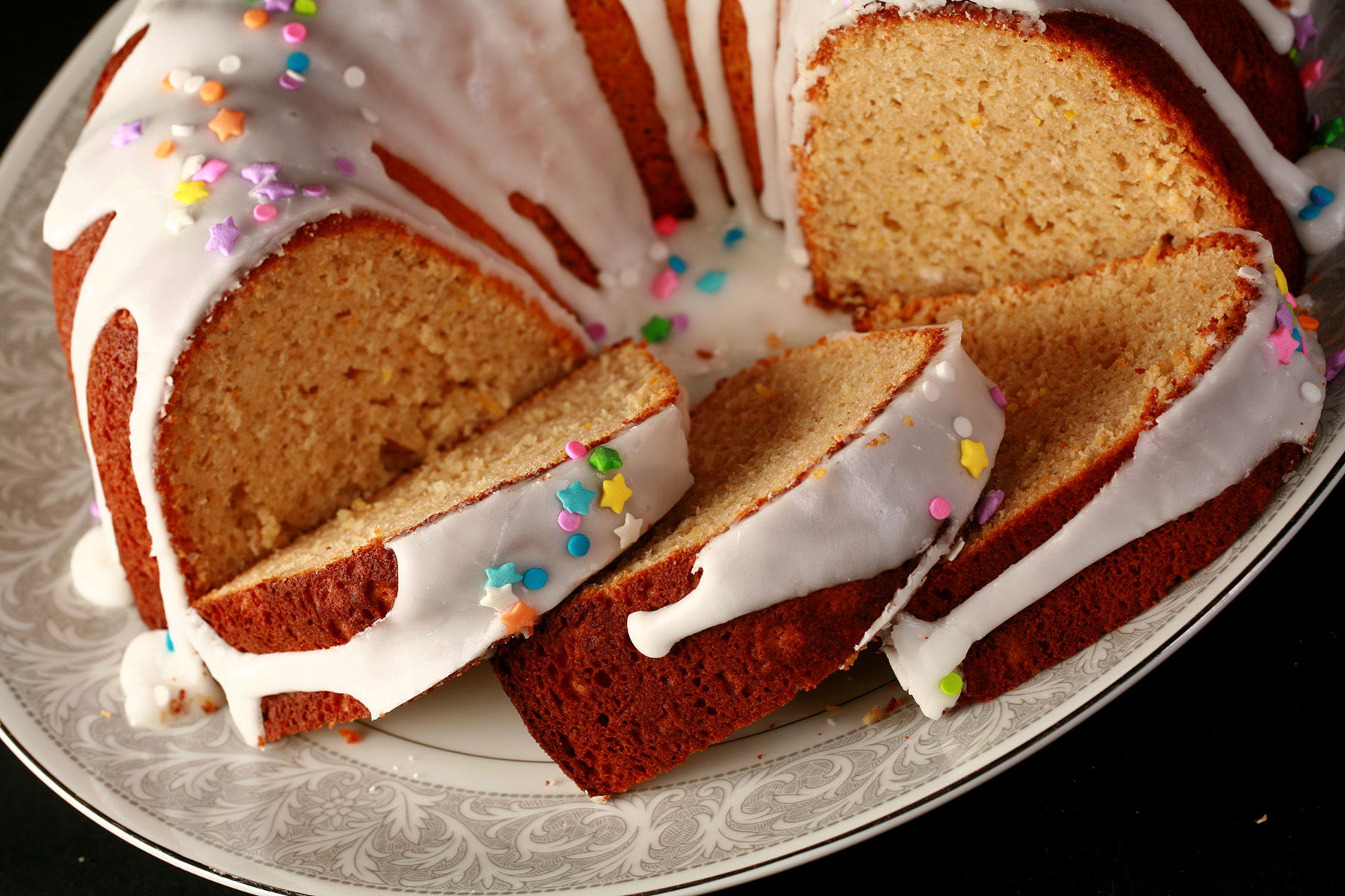 A close up view of a gluten-free paska. It's in the shape of a bundt pan, with dripping white glaze and topped with pastel coloured sprinkles. A section has been sliced, the slices are fanned out on the plate.