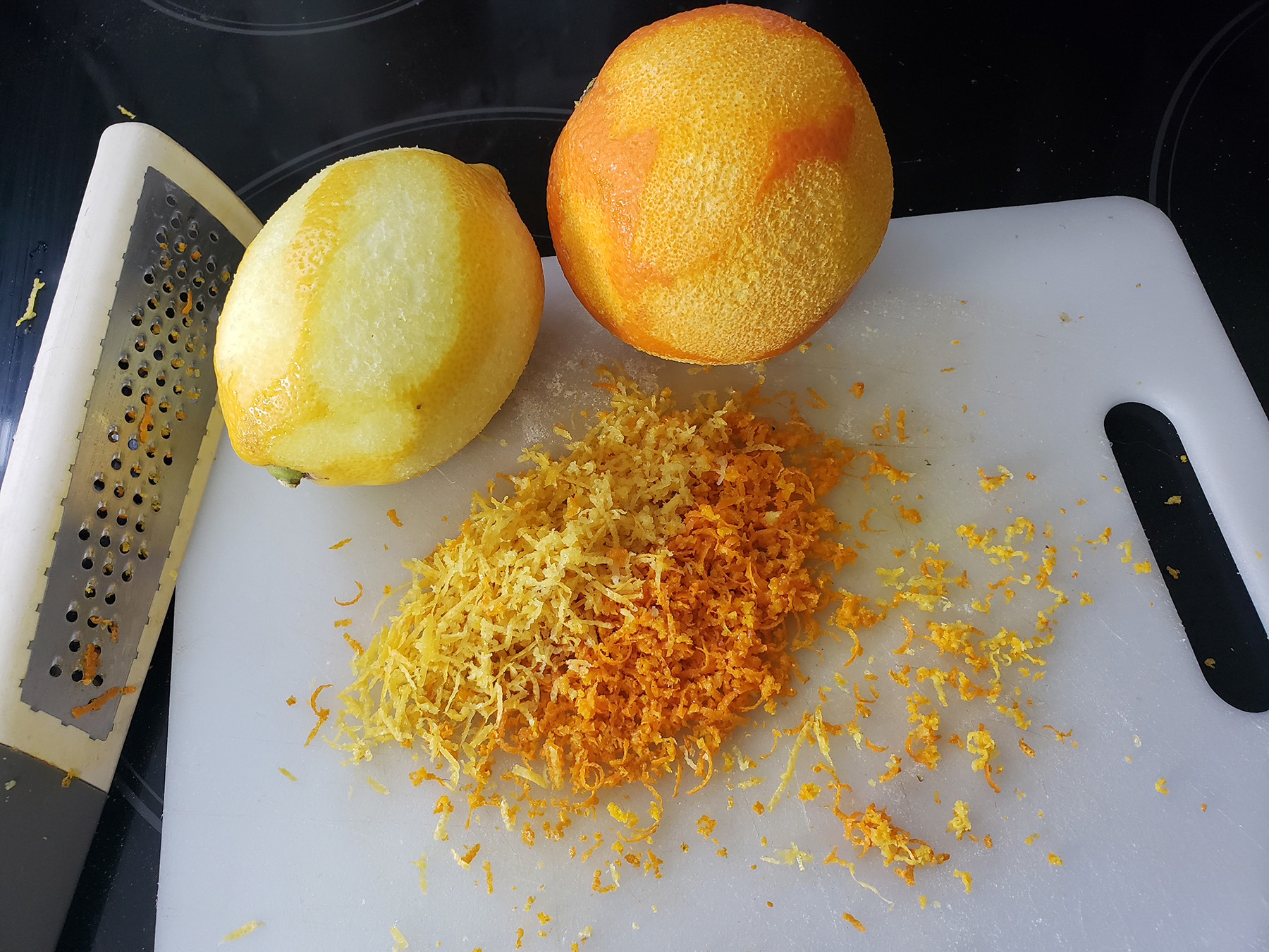 A zested orange and lemon on a white cutting board, with the zest of both displayed in piles on the cutting board.