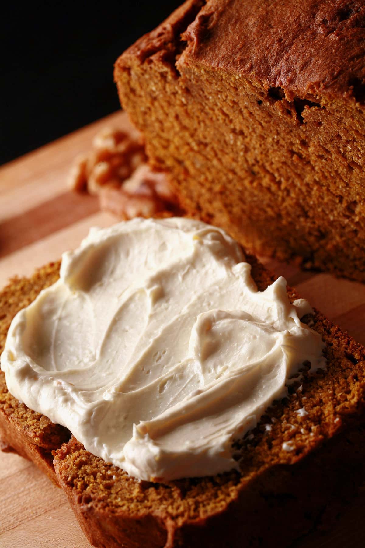 A loaf of gluten-free pumpkin bread on a cutting board, sliced. The slice is spread with cream cheese.