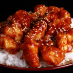 A close up view of a plate of white rice, topped with gluten-free sesame chicken. Chunks of battered and fried chicken in a glossy brown sauce, sprinkled with sesame seeds.