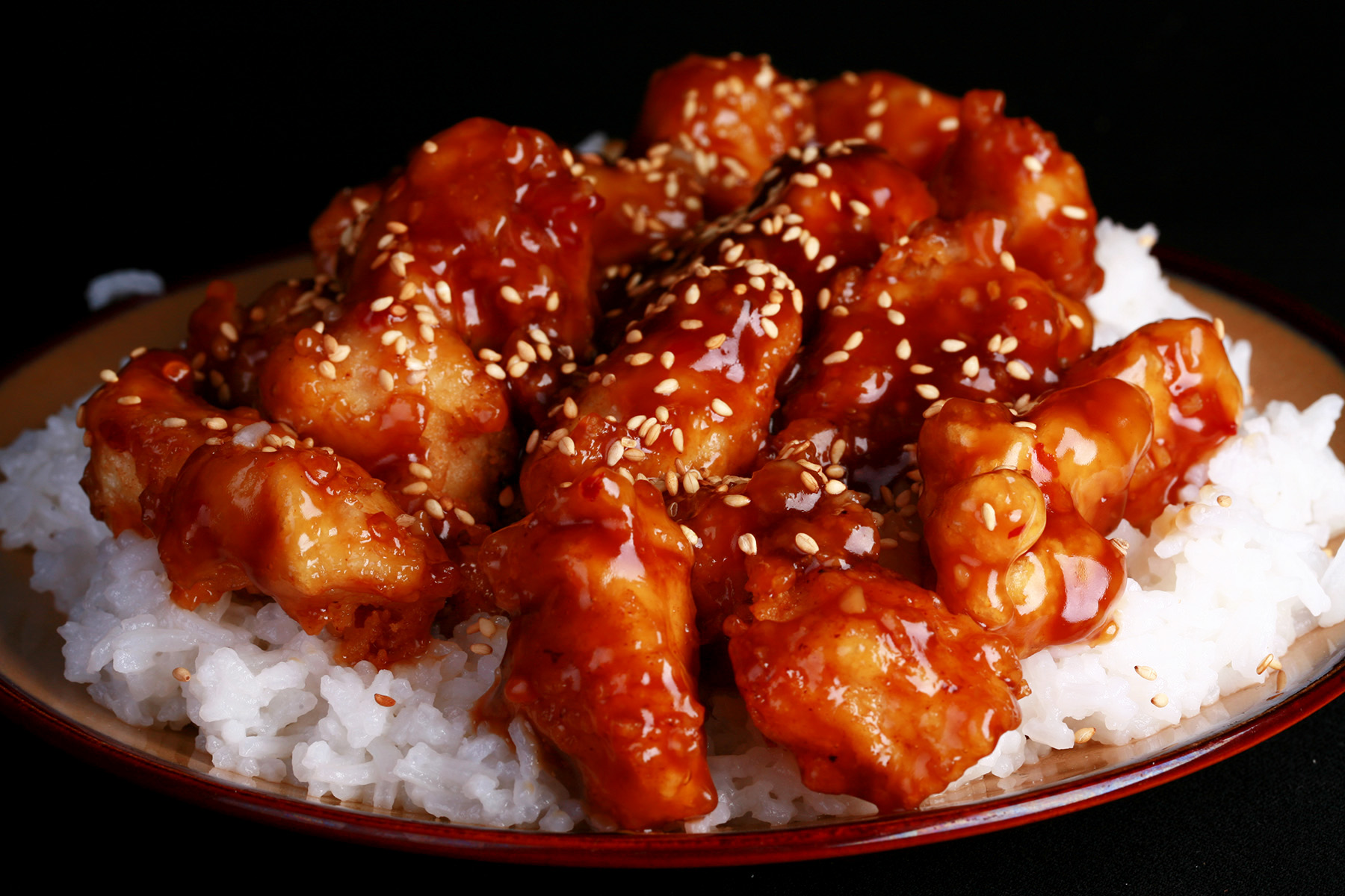 A close up view of a plate of white rice, topped with gluten-free sesame chicken. Chunks of battered and fried chicken in a glossy brown sauce, sprinkled with sesame seeds.