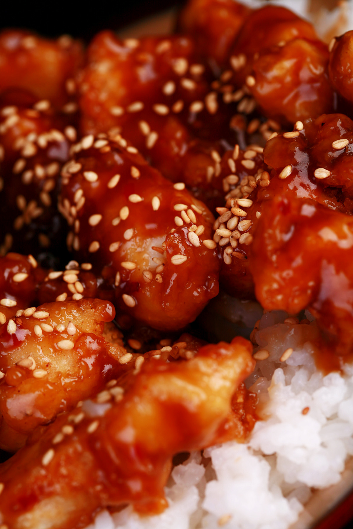 A close up view of a plate of white rice, topped with gluten-free sesame chicken. Chunks of battered and fried chicken in a glossy brown sauce, sprinkled with sesame seeds.