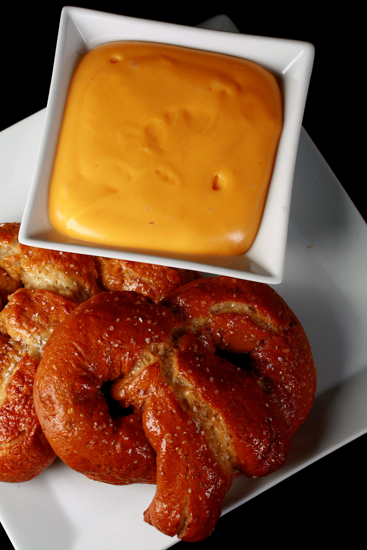2 soft gluten-free pretzels on a plate, beside a bowl of mustard-cheddar dip.