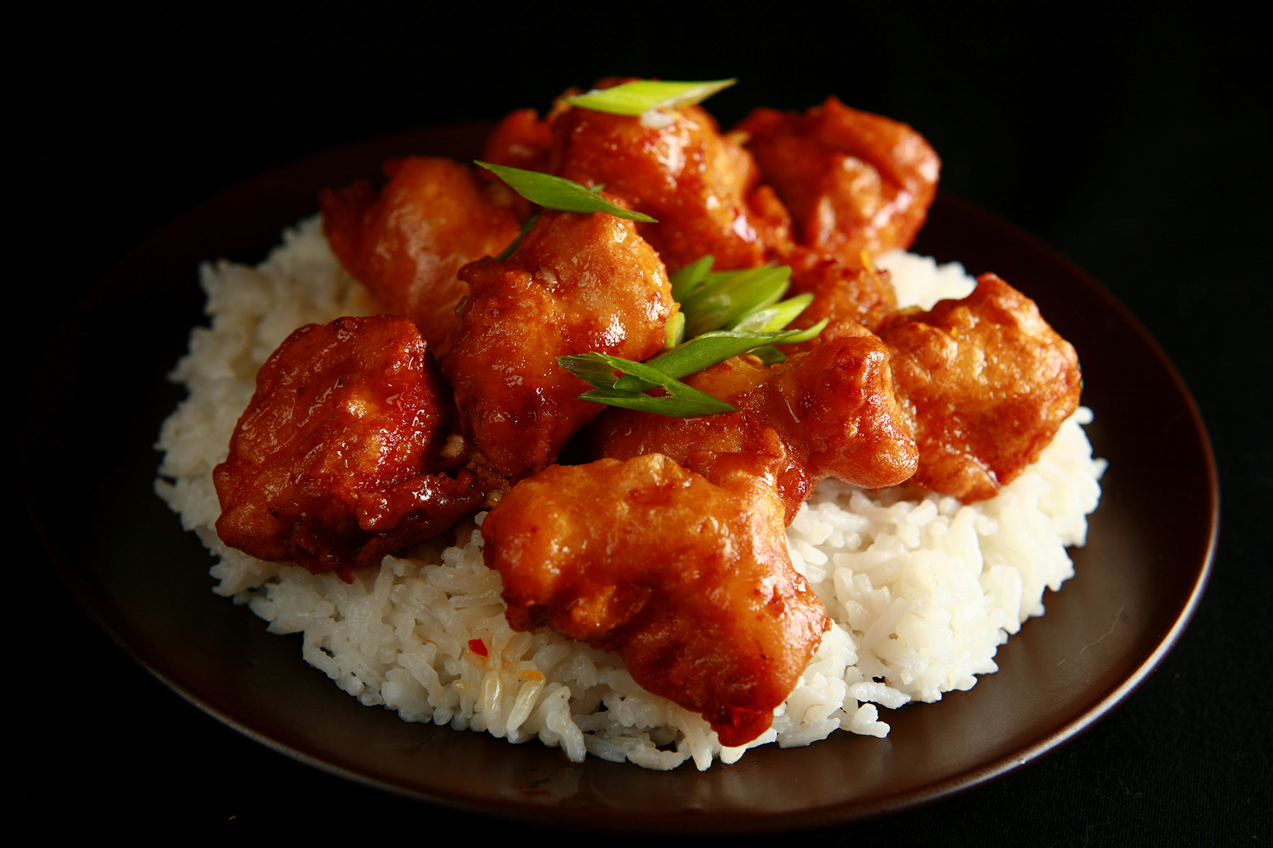 A plate of rice, covered with gluten free orange chicken: Chunks of battered and deep fried chicken coated in a glossy orange sauce, garnished with sliced green onions.