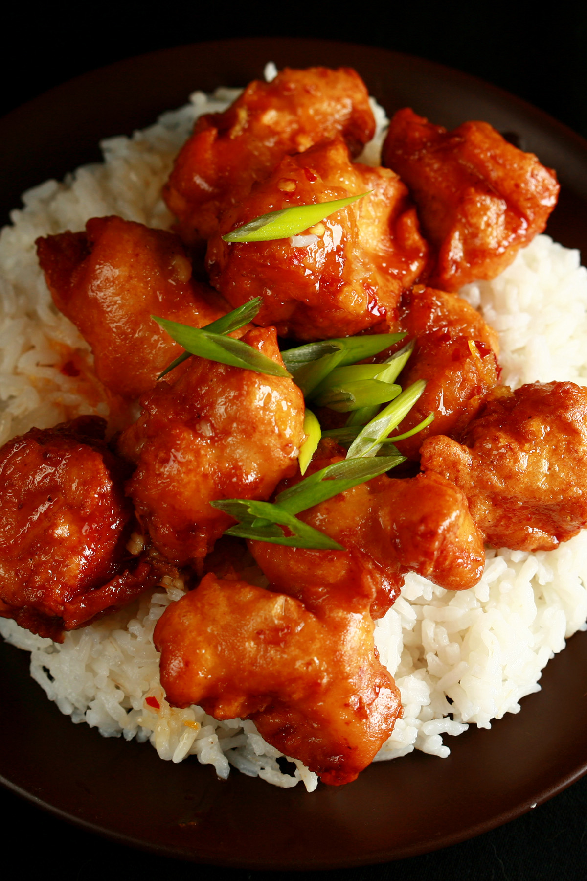 A plate of rice, covered with gluten-free orange chicken: Chunks of battered and deep fried chicken coated in a glossy orange sauce, garnished with sliced green onions.