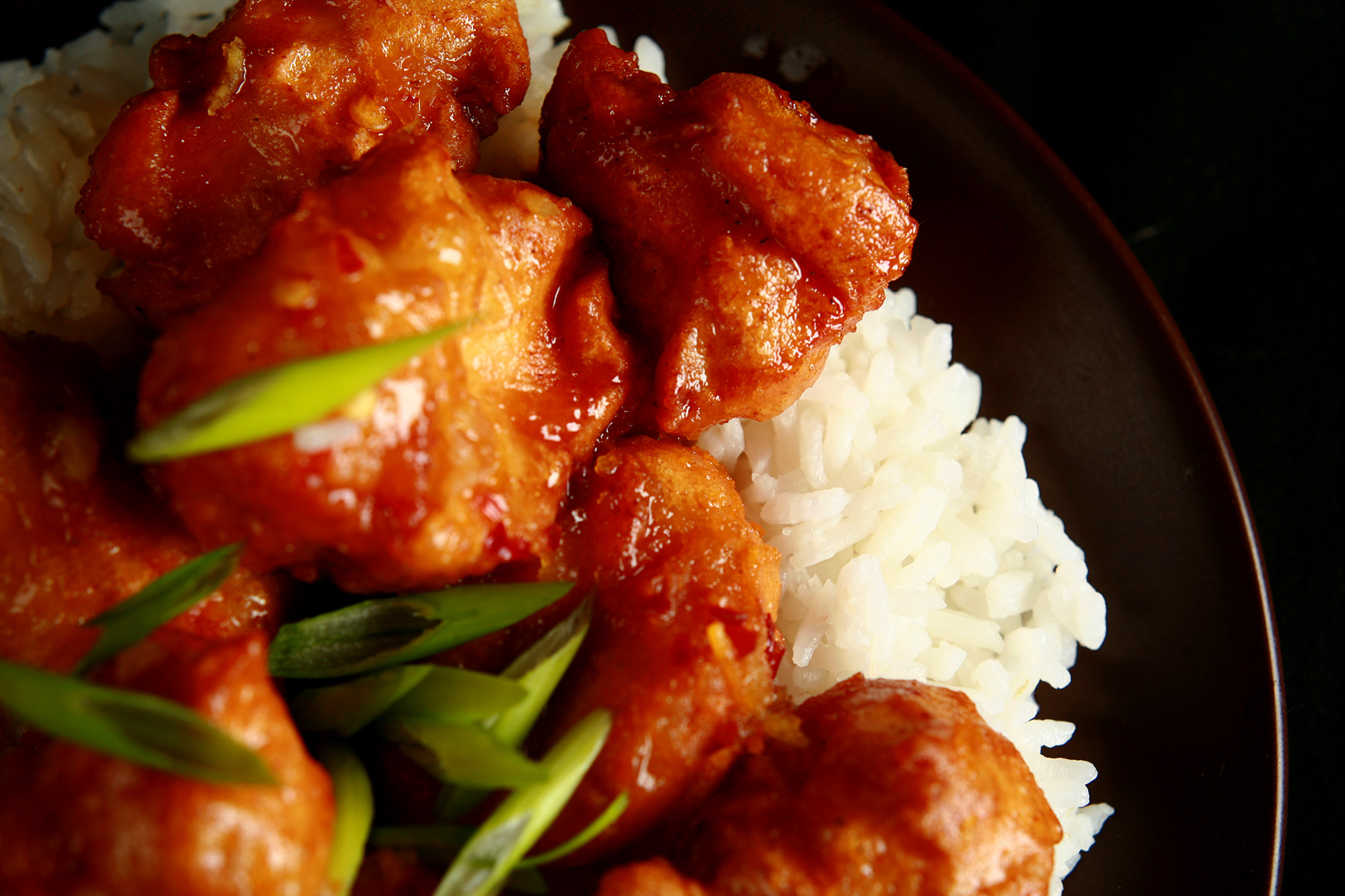 A plate of rice, covered with spicy orange chicken: Chunks of battered and deep fried chicken coated in a glossy orange sauce, garnished with sliced green onions.