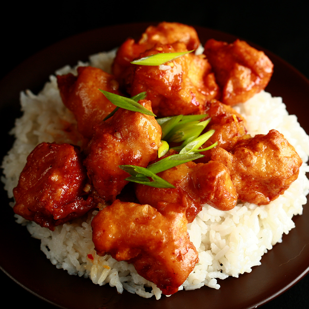 A plate of rice, covered with gluten-free spicy orange chicken: Chunks of battered and deep fried chicken coated in a glossy orange sauce, garnished with sliced green onions