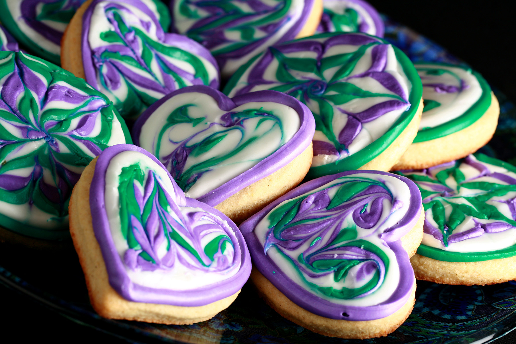Close up view of a plate of heart shaped gluten-free sugar cookies. They are decorated with swirls of teal and lavender over a base of white frosting.
