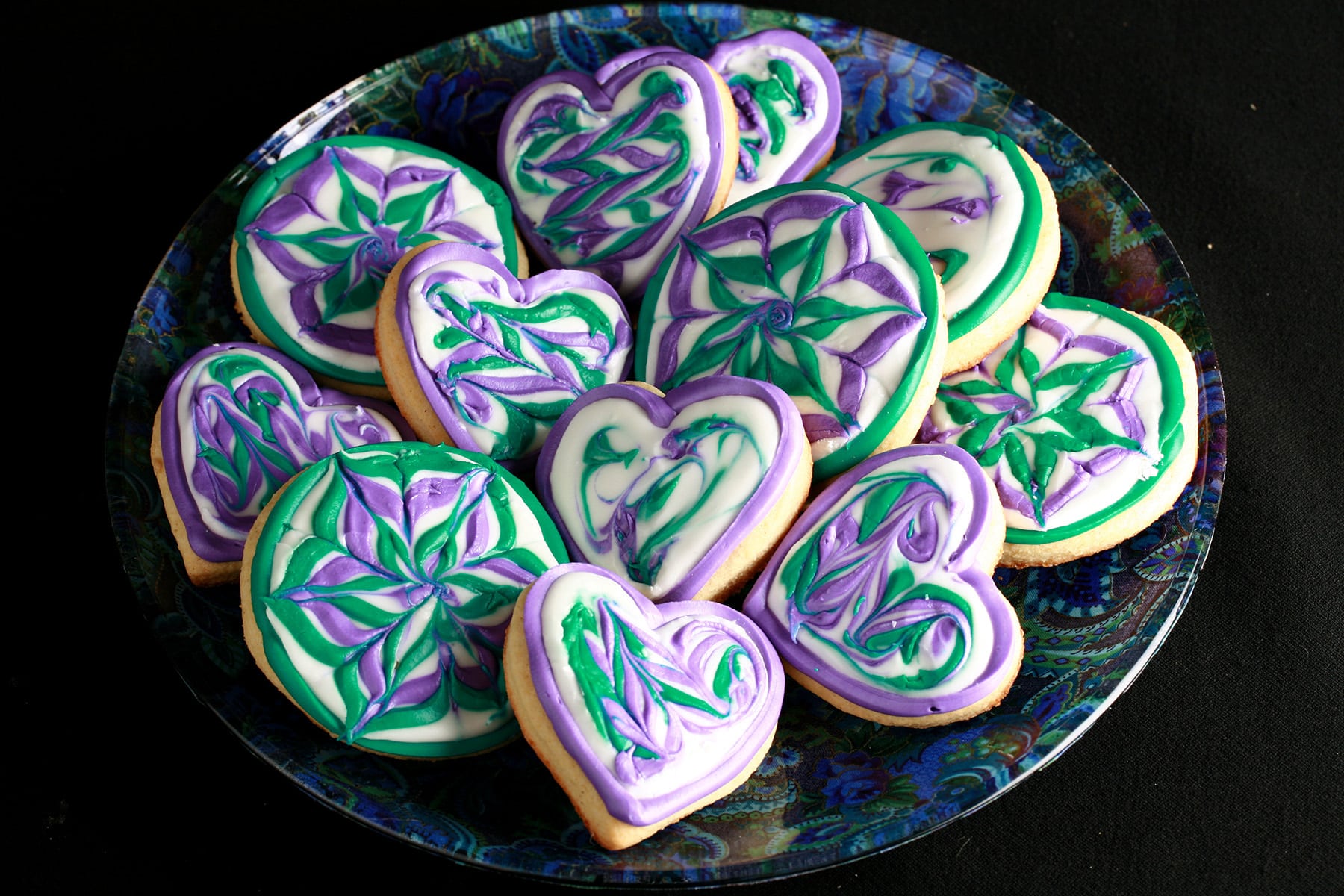 Close up view of a plate of heart shaped gluten-free sugar cookies. They are decorated with swirls of teal and lavender over a base of white frosting.