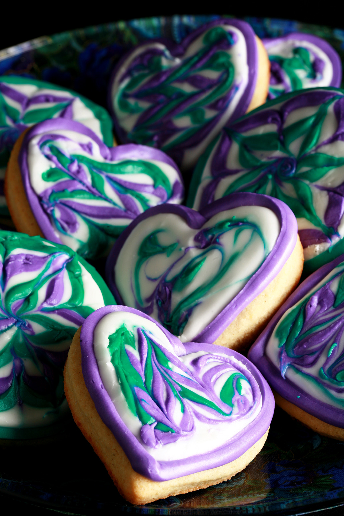 Close up view of a plate of heart shaped cookies decorated with swirls of teal and lavender over white.