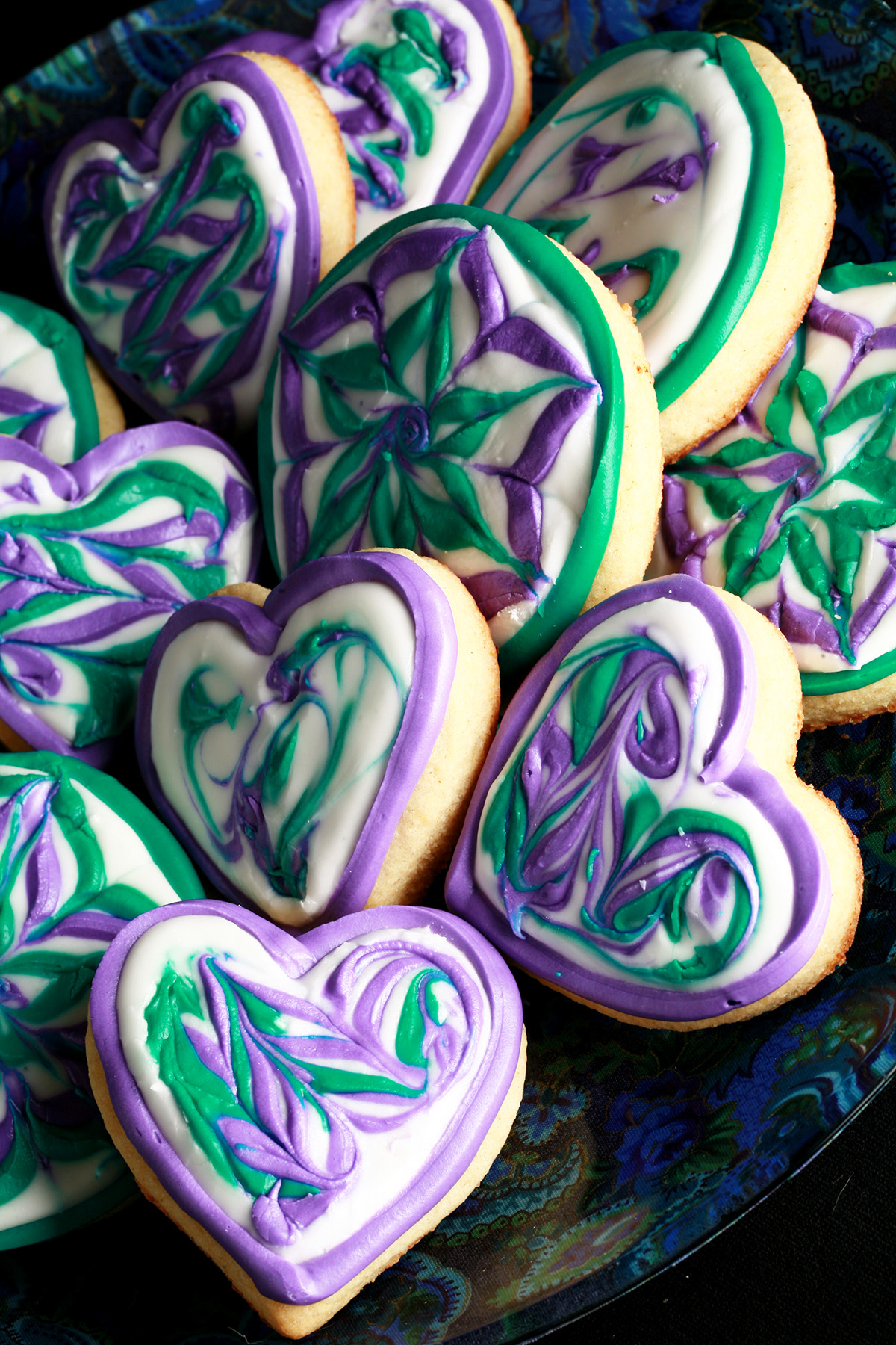 Close up view of a plate of heart shaped cookies decorated with swirls of teal and lavender over white.