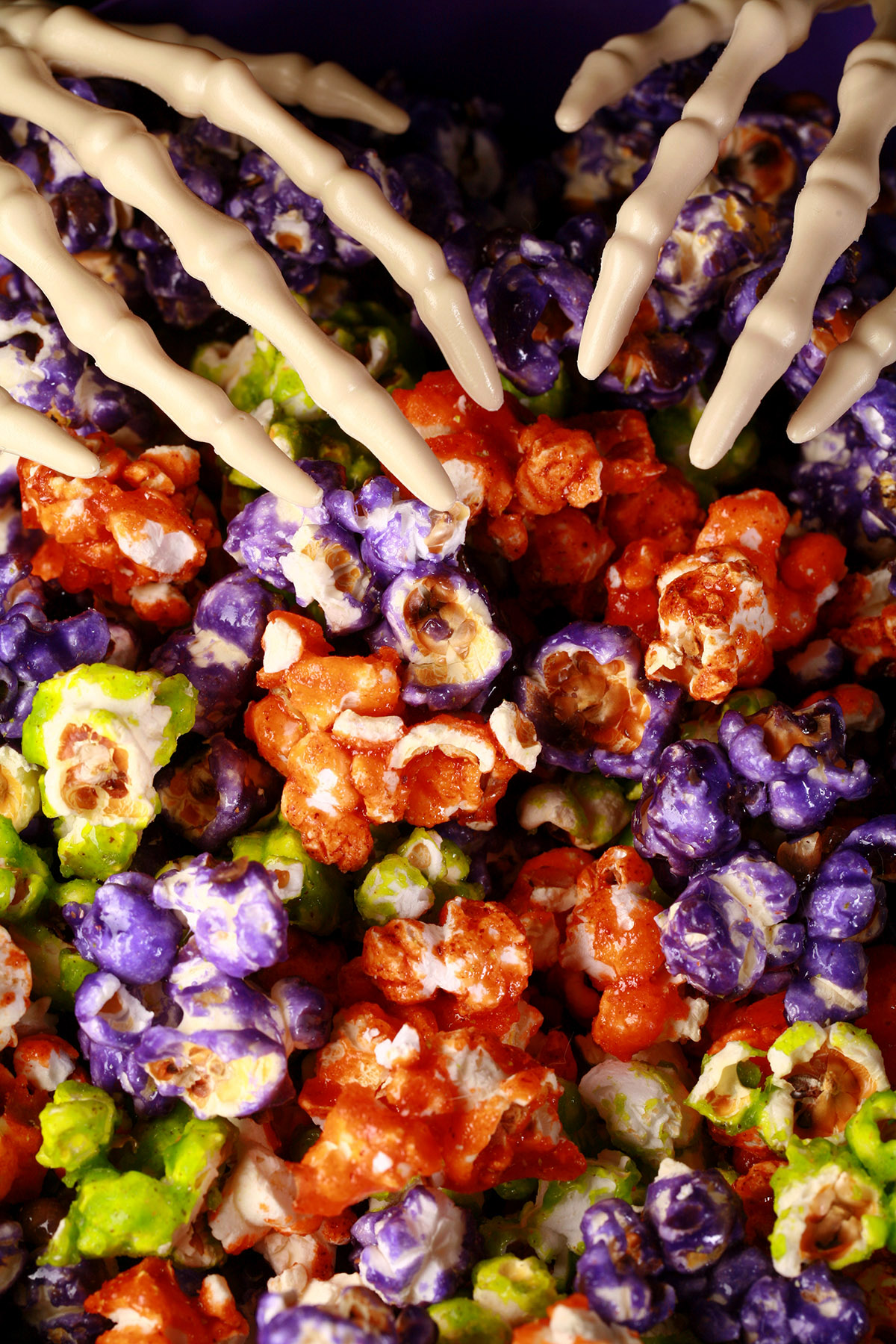 A bowl of Halloween popcorn - orange, purple, and lime green glazed popcorn, mixed together. A pair of skeleton hand serving spoons are visible.