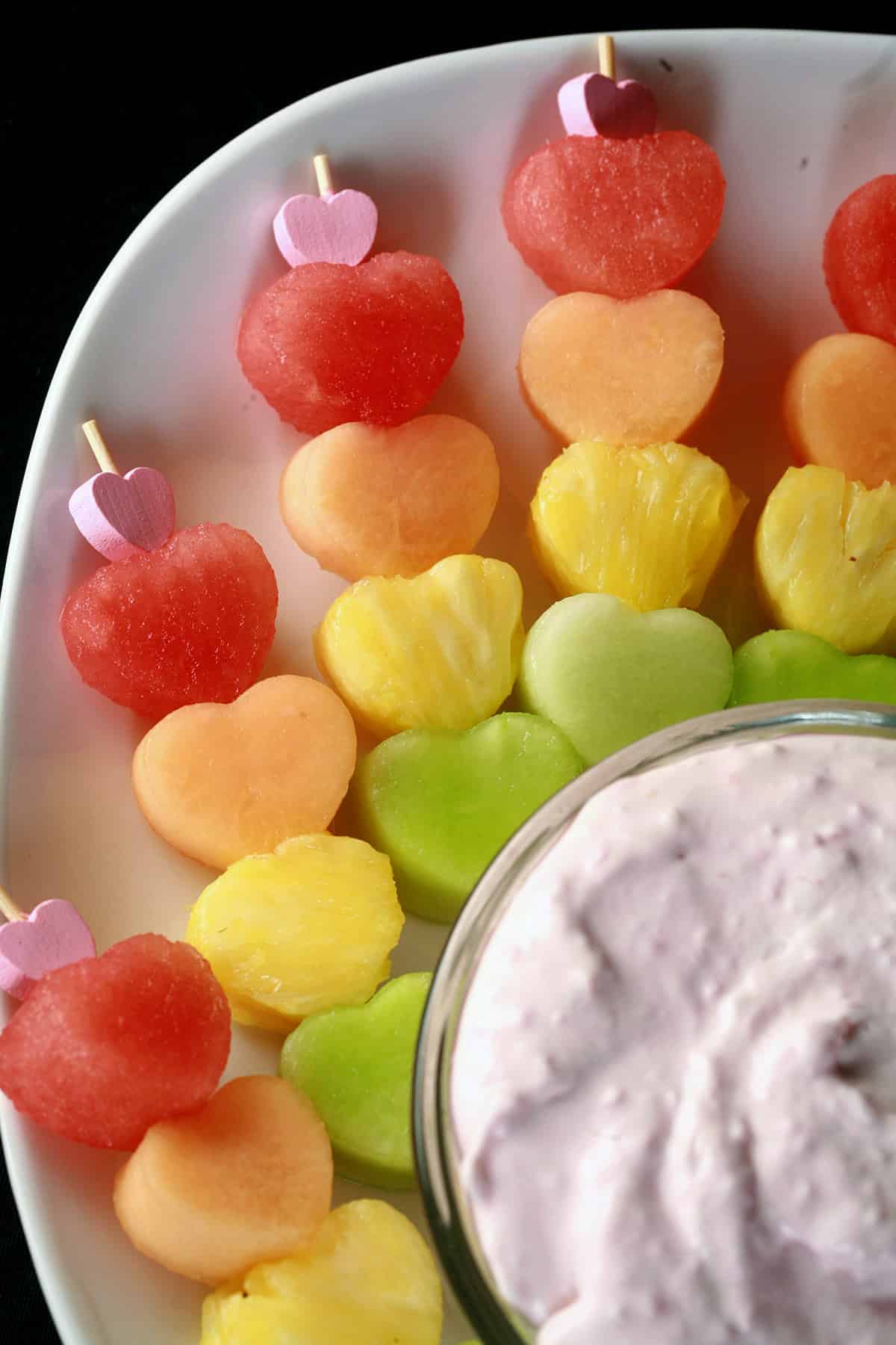 Several skewers of heart shaped fruit - watermelon, cantaloupe, pineapple, and honeydew - next to a bowl of raspberry cheesecake dip.