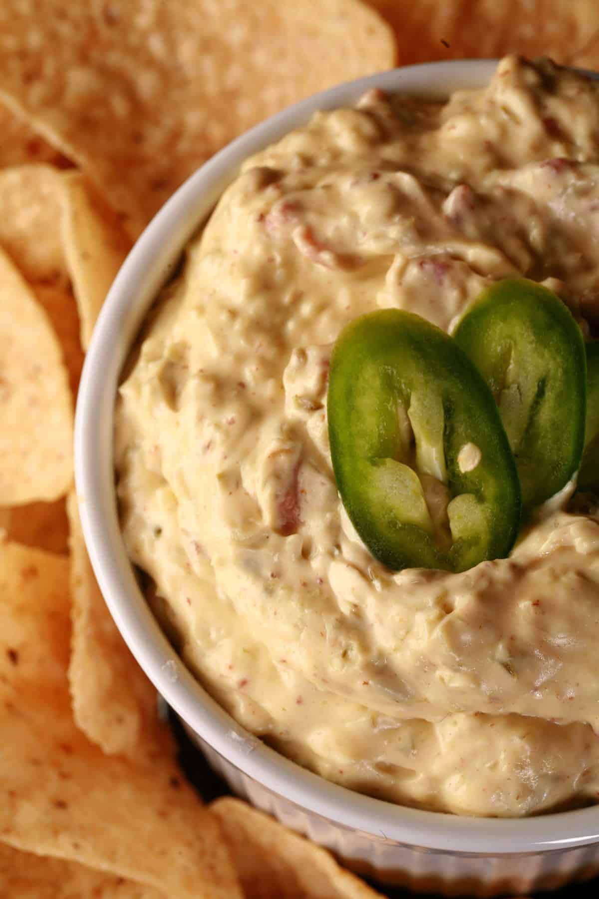 A ramekin of bacon jalapeno dip, surrounded by corn chips and topped with sliced jalapenos.