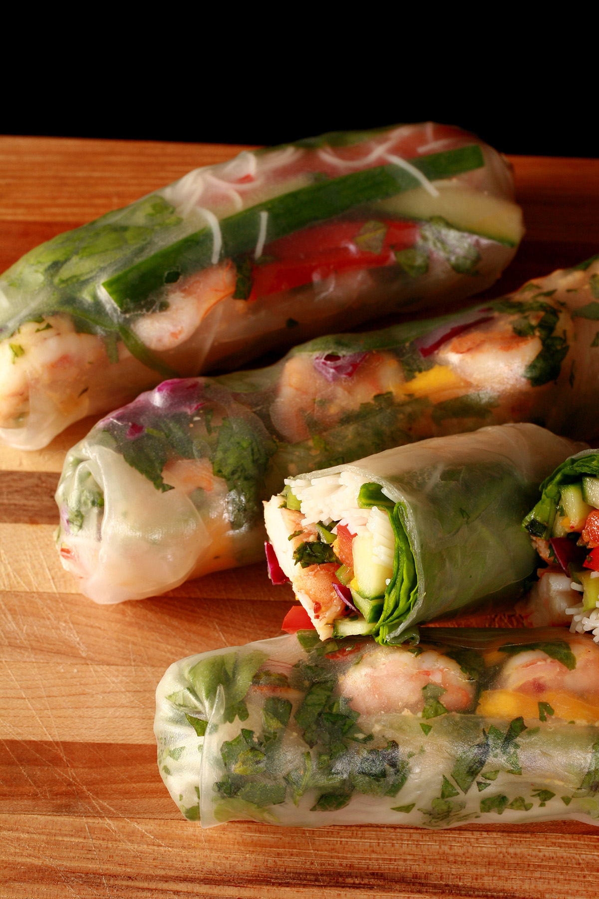 Several mango-shrimp spring rolls on a cutting board, along with a small bowl of peanut sauce.