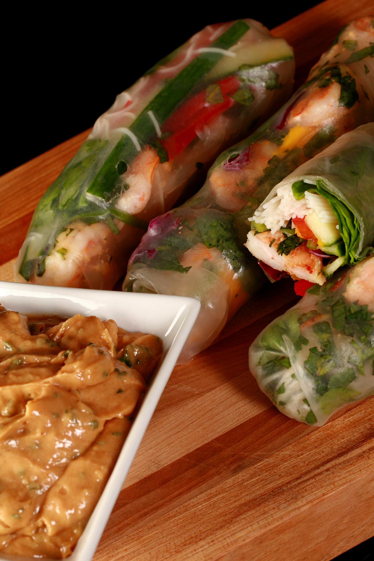Several mango-shrimp spring rolls on a cutting board, along with a small bowl of peanut sauce.