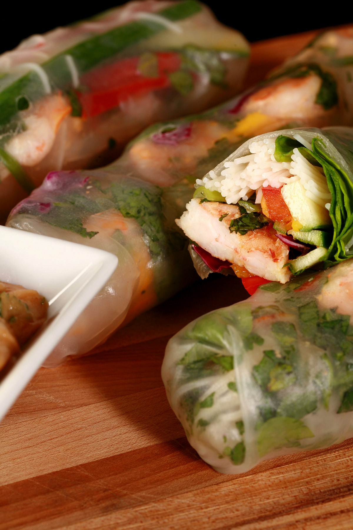 Several mango-shrimp spring rolls on a cutting board, along with a small bowl of peanut sauce.