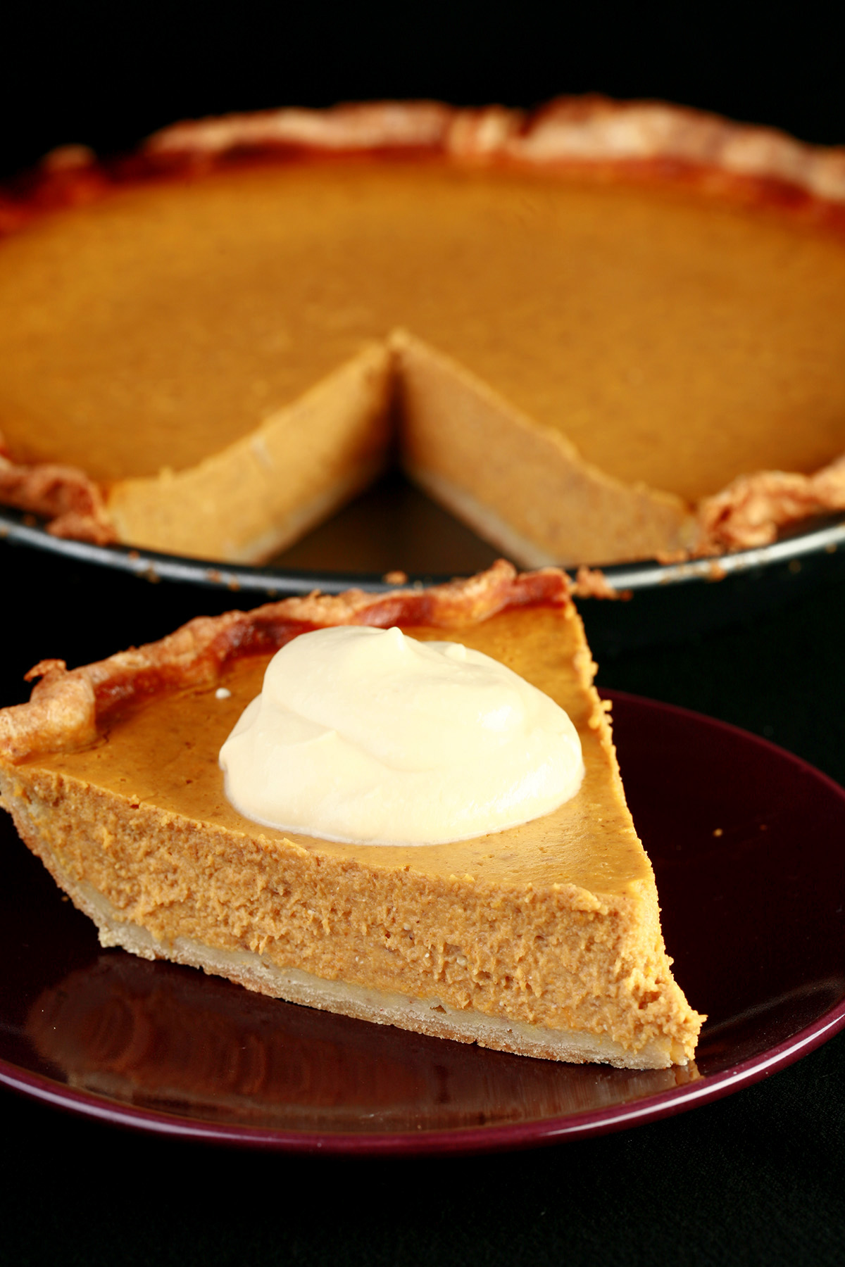 A slice of gluten-free maple pumpkin pie with maple cream, in front of a whole pumpkin pie.