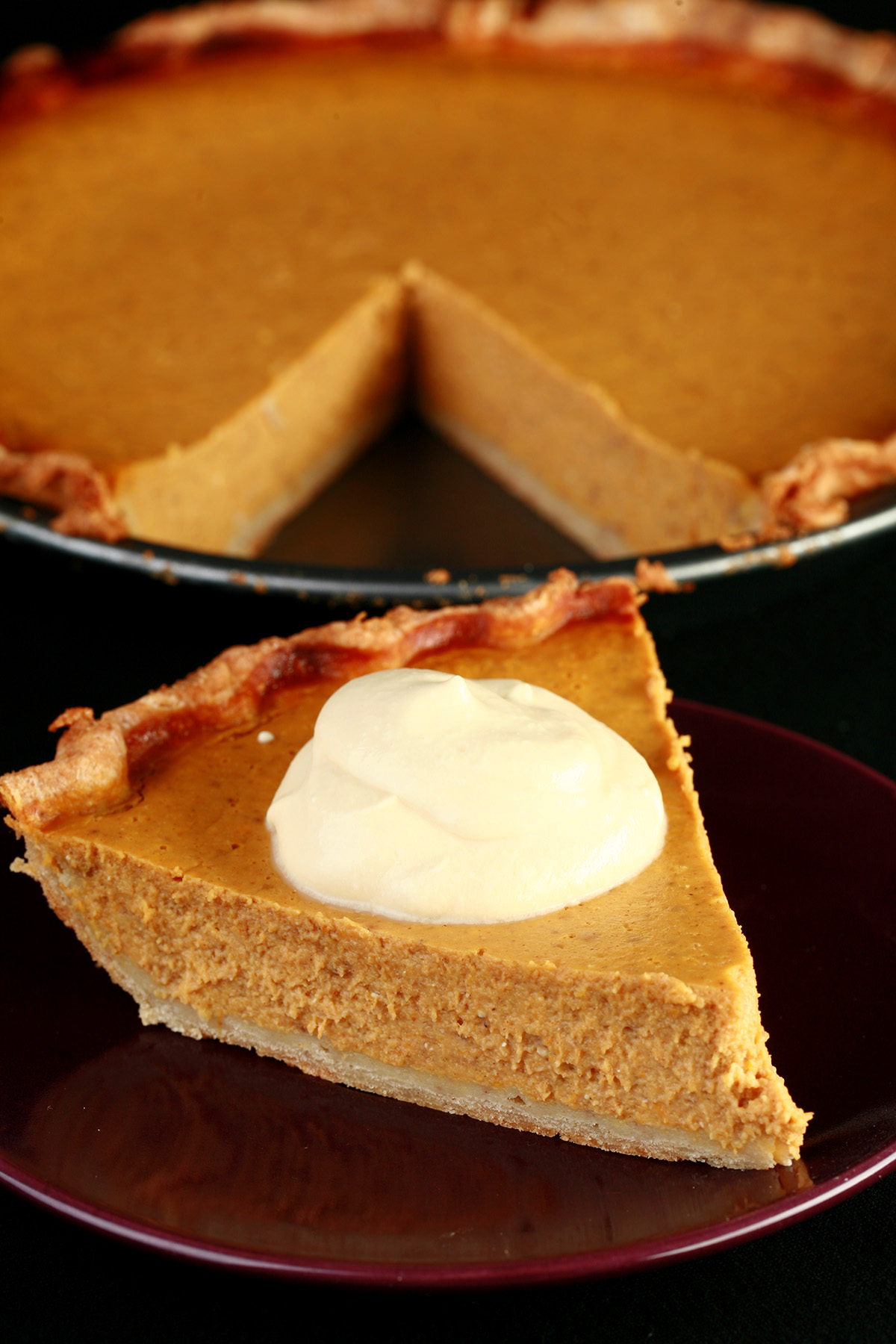 A slice of pumpkin pie with maple cream, in front of a whole pumpkin pie.