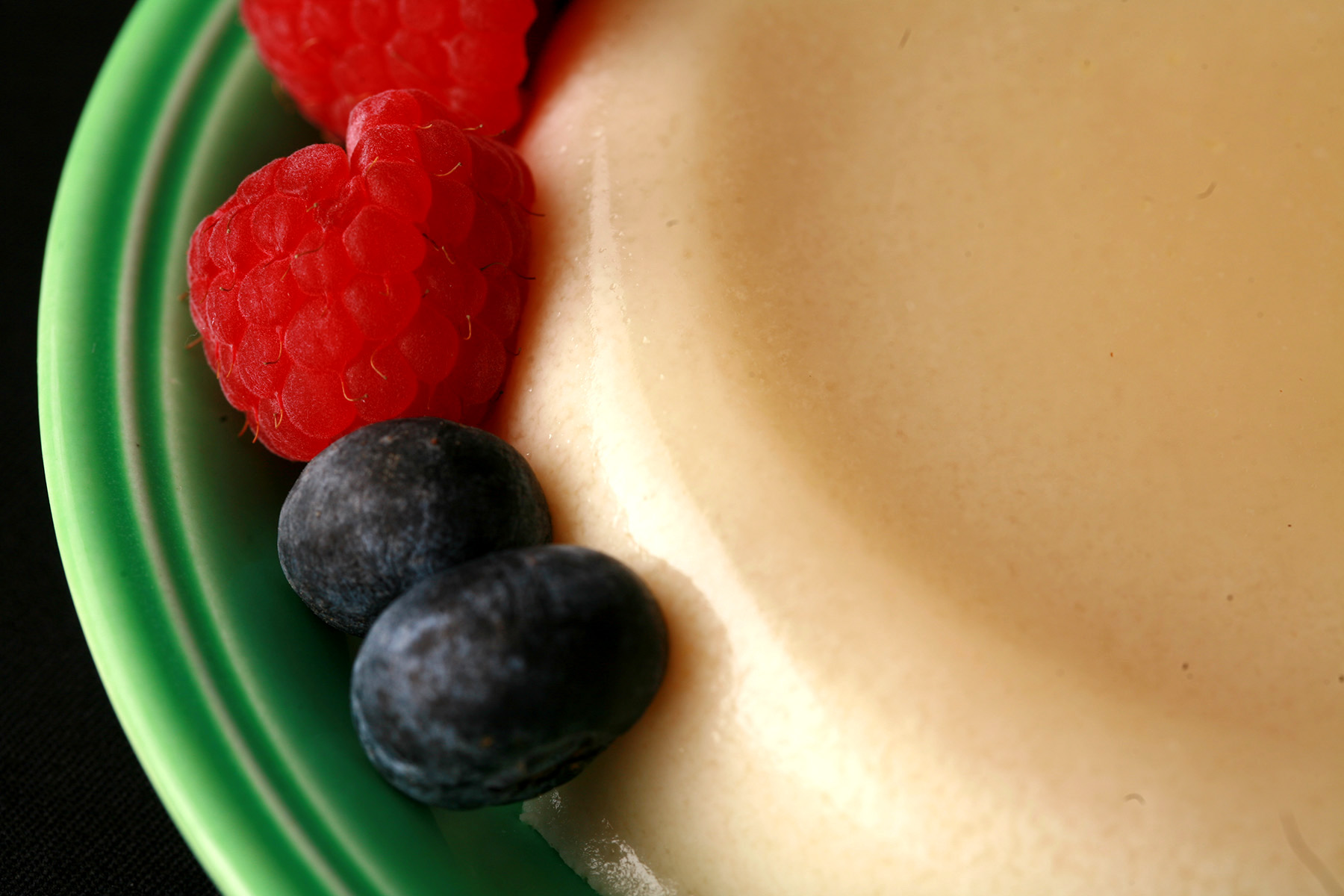 A round serving of calamansi AIP panna cotta on a green plate, with raspberries and blueberries next to it.