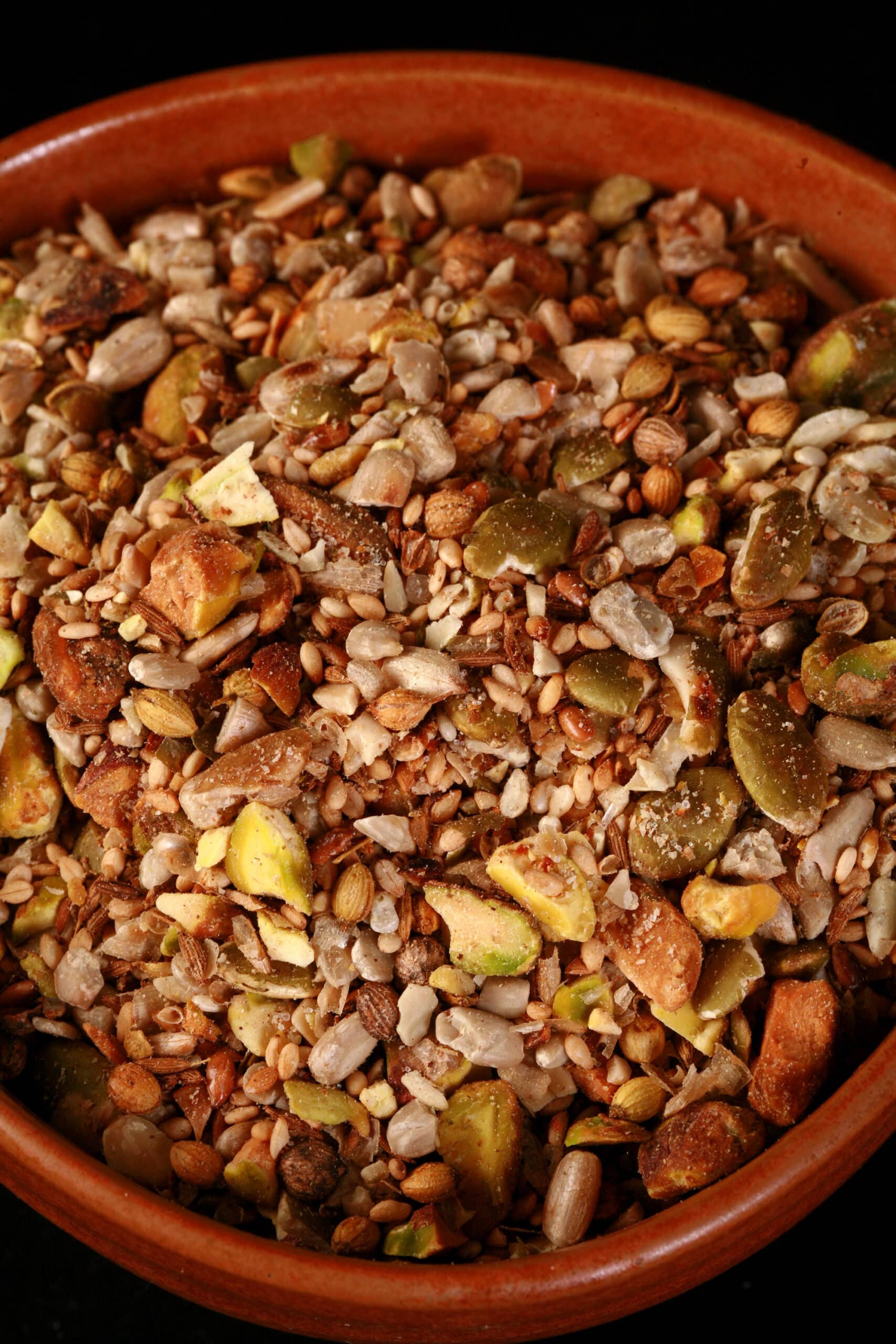 A small brown bowl of pistachio dukkag spice mix. Chunks of pistachios, coriander, sunflower seeds, and pumpkin seeds are visible.