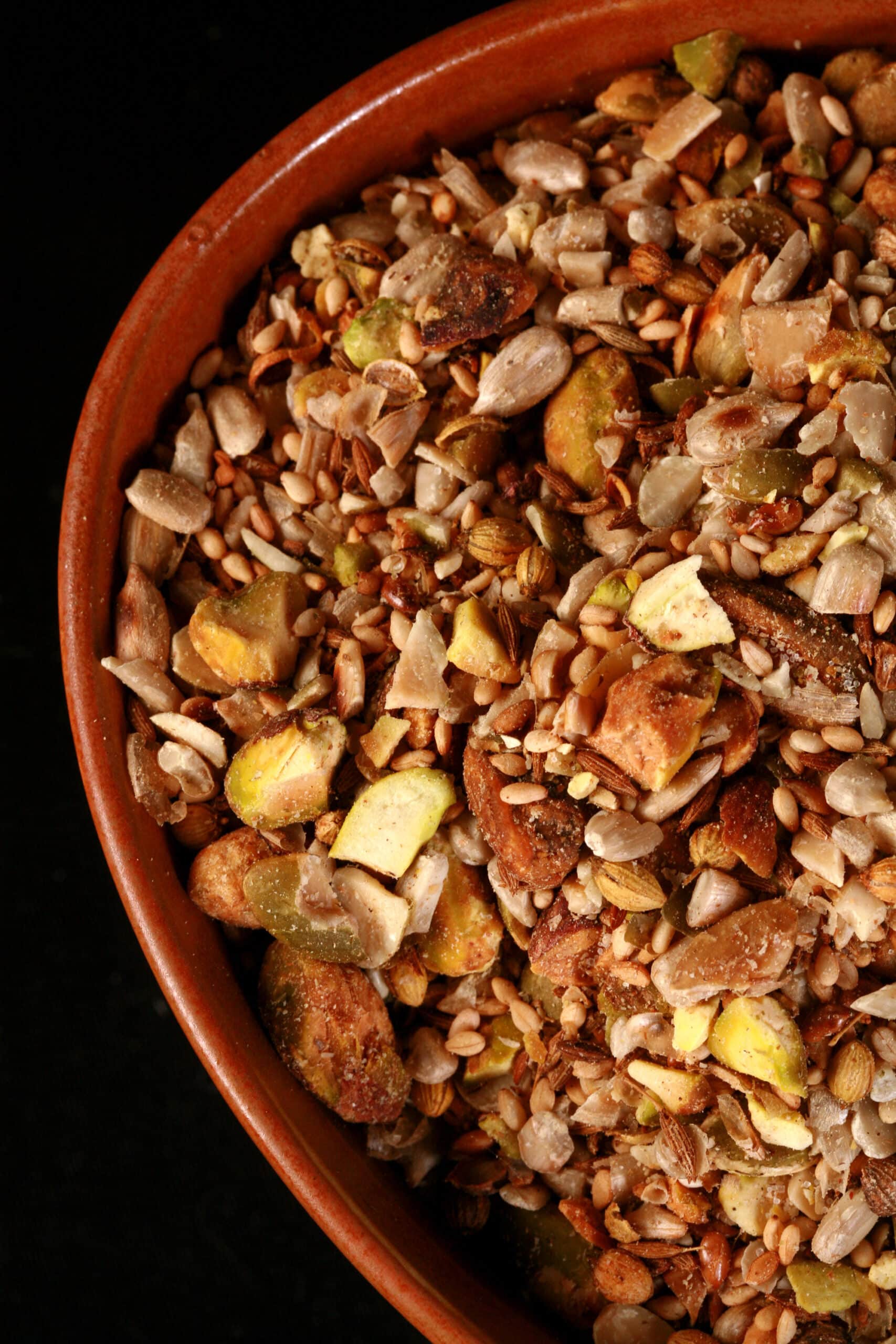 A small brown bowl of pistachio dukkag spice mix. Chunks of pistachios, coriander, sunflower seeds, and pumpkin seeds are visible.