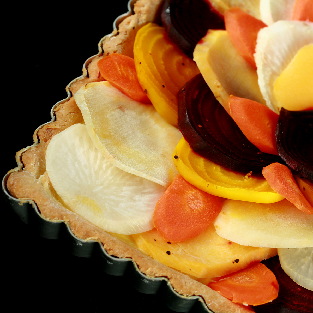 A close up view of a square root tart - a tart cooked in a square pan, with slices of a variety of root veggies arranged on top.