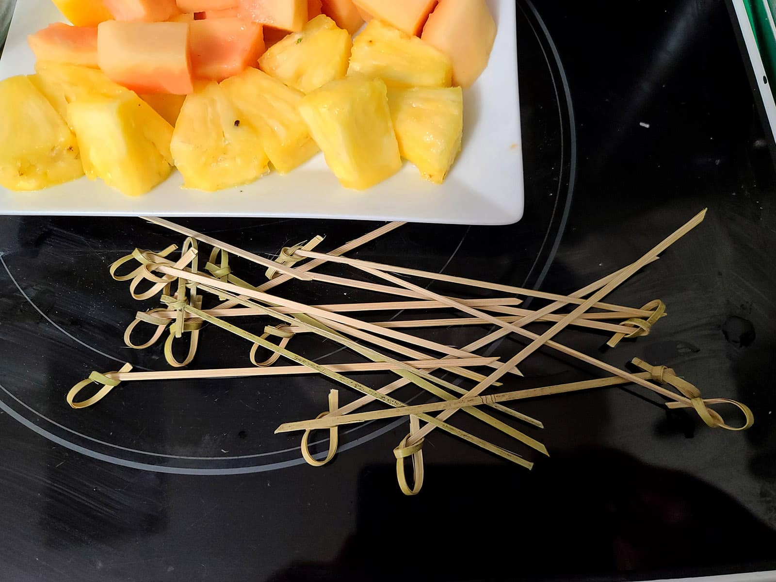 A pile of bamboo skewers in front of a plate of tropical fruit chunks.