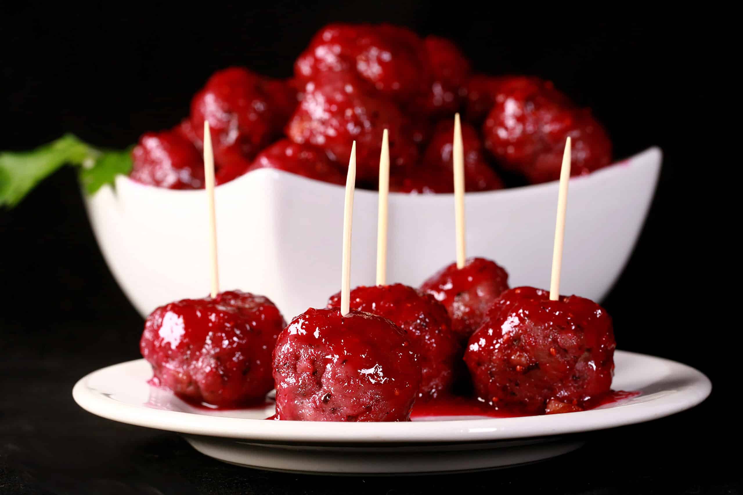 A bowl of gluten free turkey meatballs in a bright red cranberry glaze. There are several meatballs with toothpicks on them on a plate in front of the bowl.